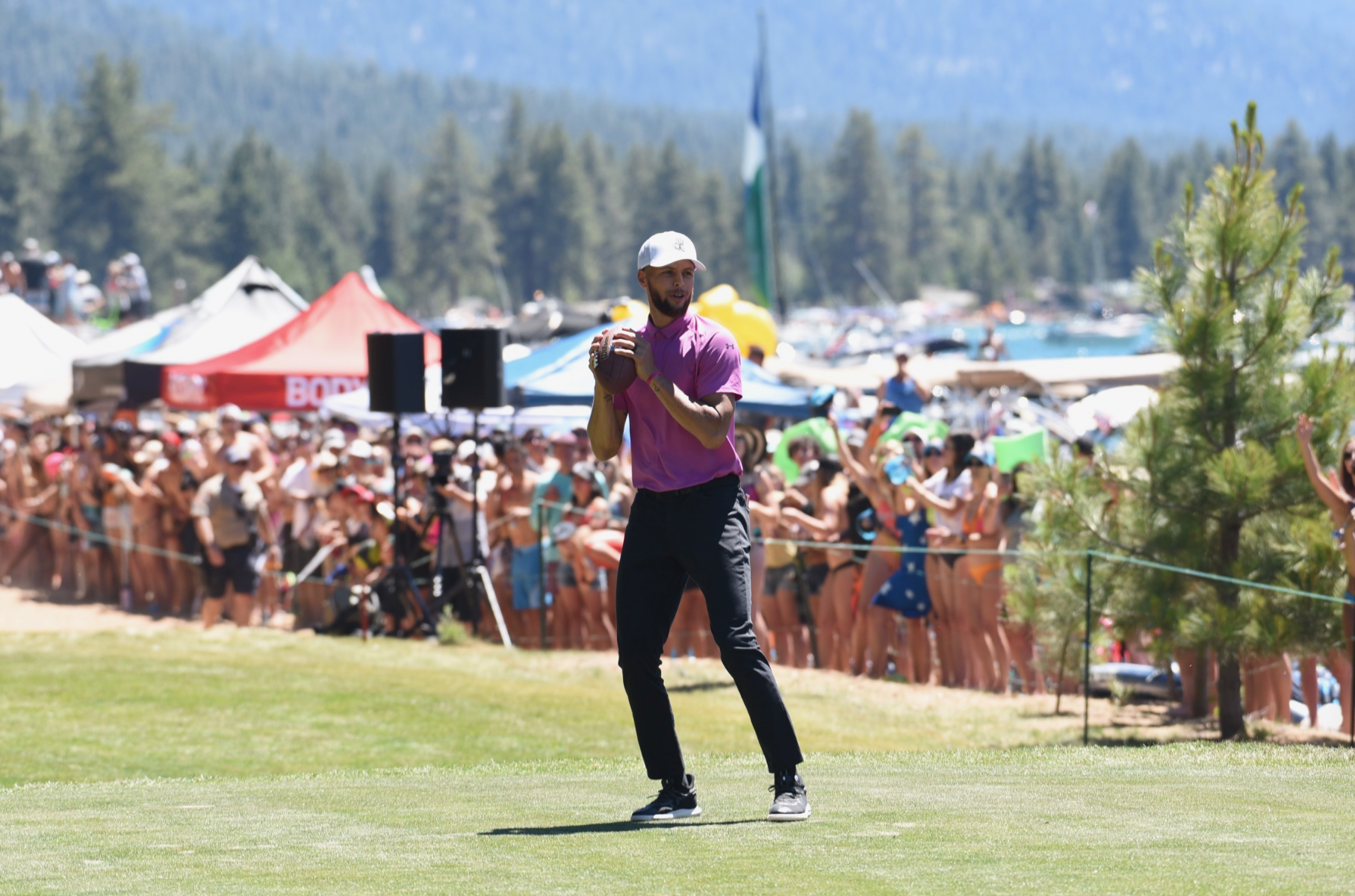 A person wearing a purple shirt and black pants is preparing to throw a football on a grassy field, surrounded by the best things to do in Tahoe in July. A crowd of people, tents, and forested mountains fill the background, highlighting the outdoor event on this sunny day.